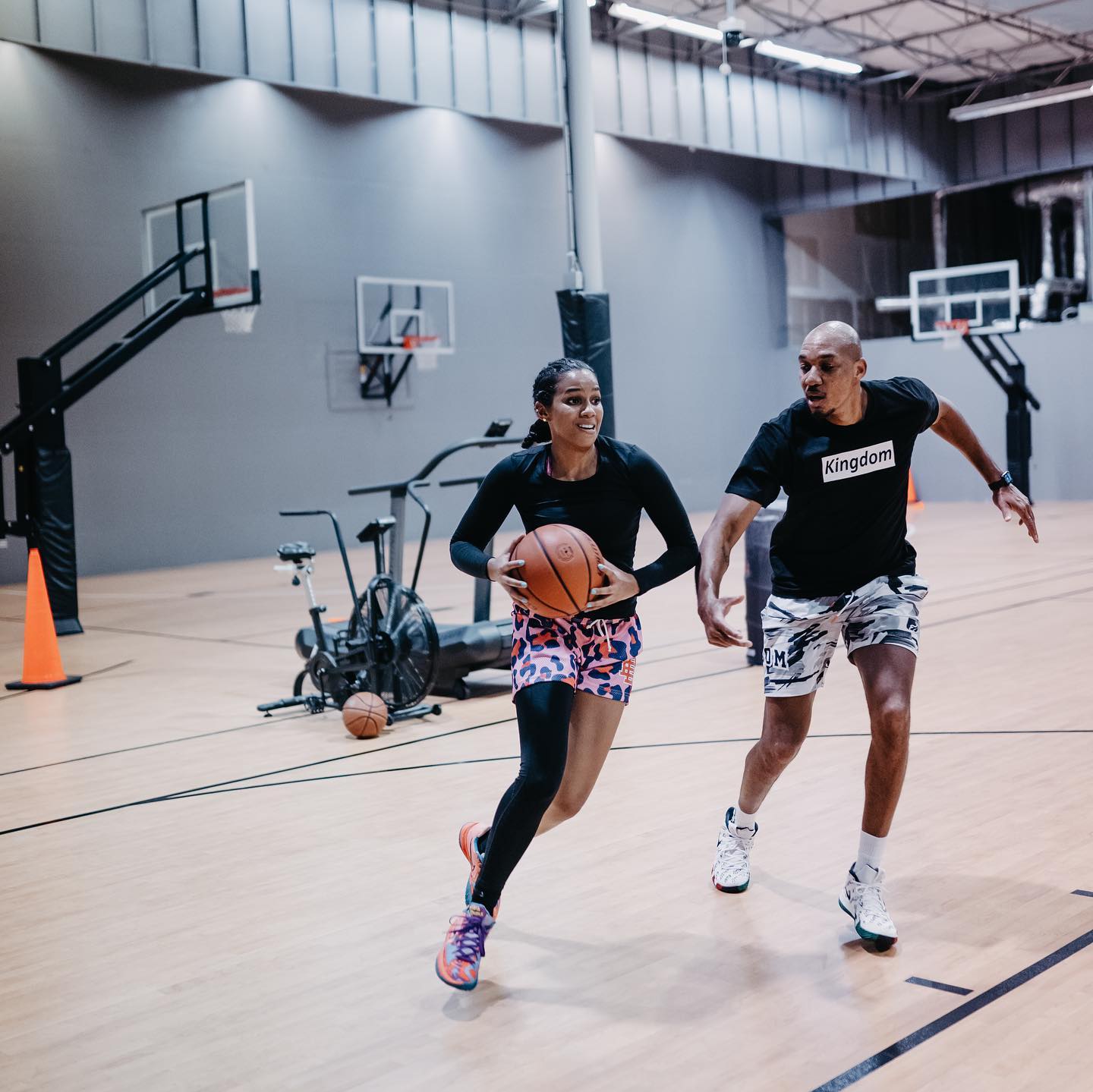 Woman dribbling basketball, chased by a man in a gym. Both are smiling; basketball court setting.