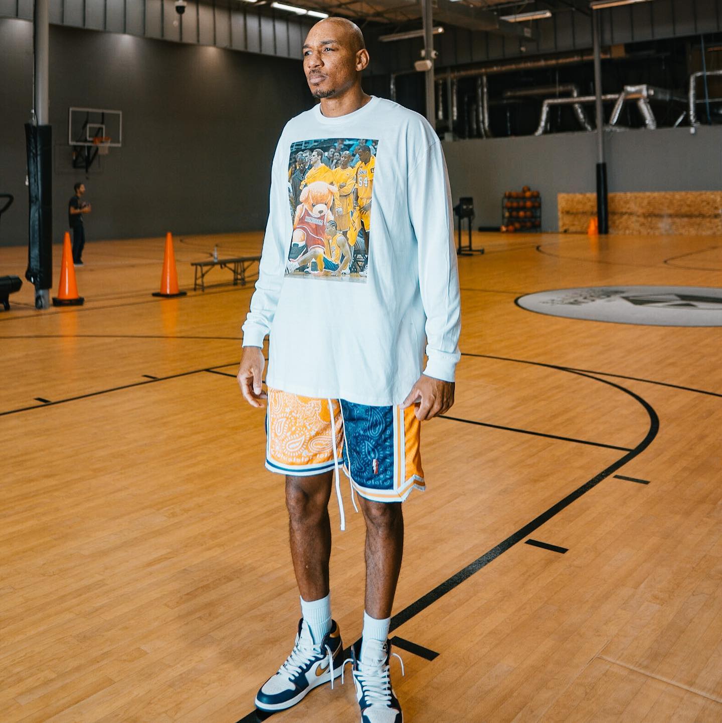 Man standing on a basketball court wearing a long-sleeve shirt and shorts; sporting blue and white high-top sneakers.
