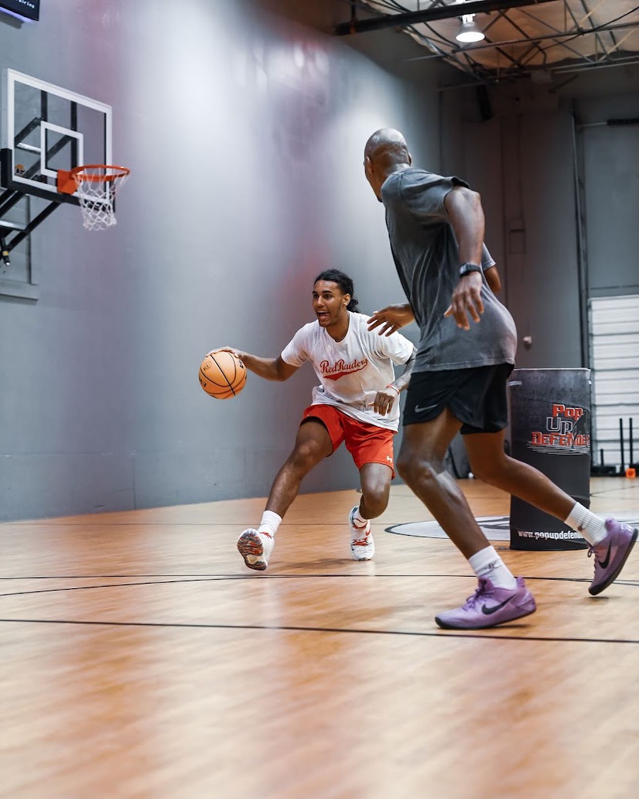 Two basketball players practicing in a gym. One dribbles, the other guards. Wooden floor, gray walls.