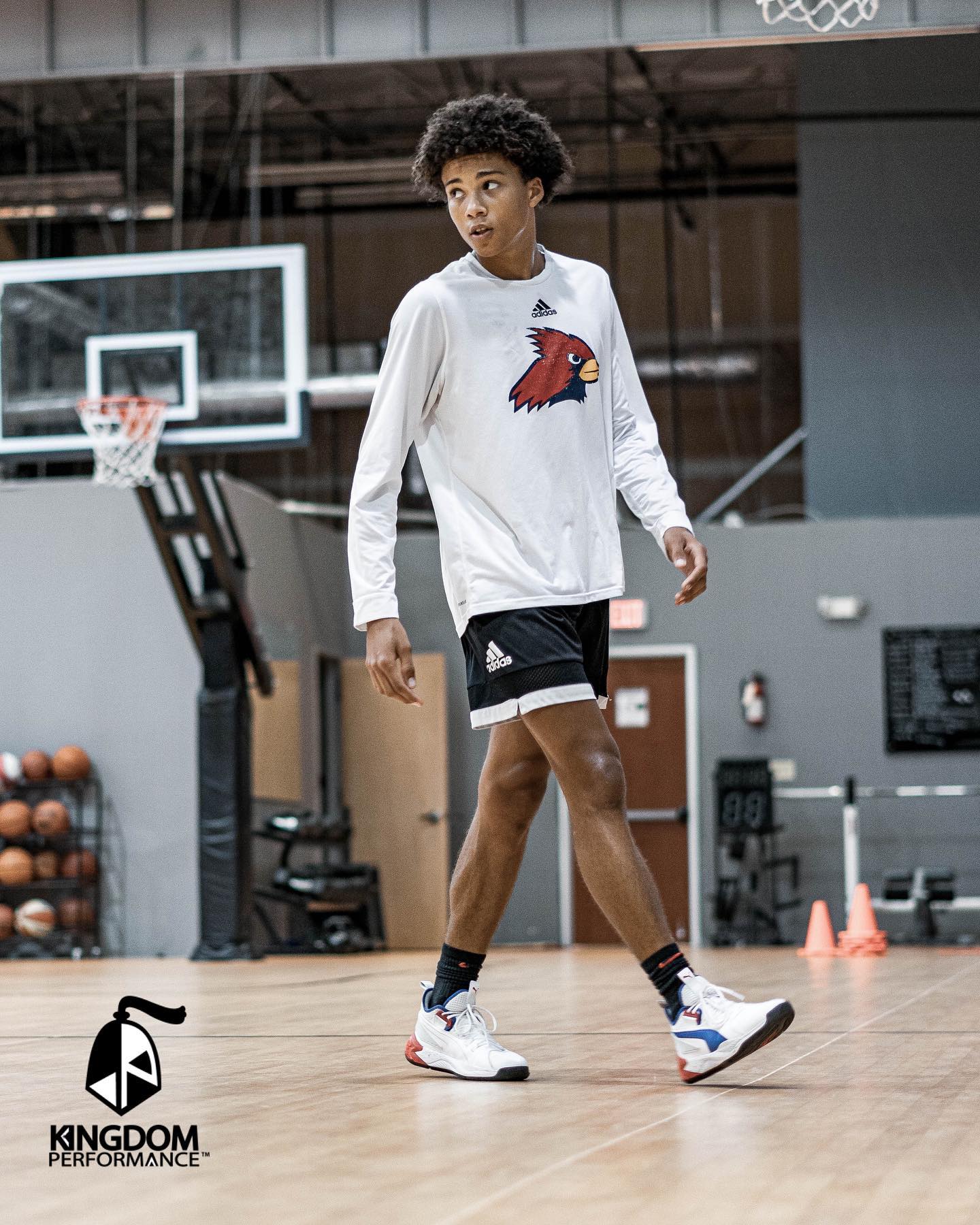 Young basketball player in gym, wearing white long-sleeve shirt, black shorts, and sneakers.