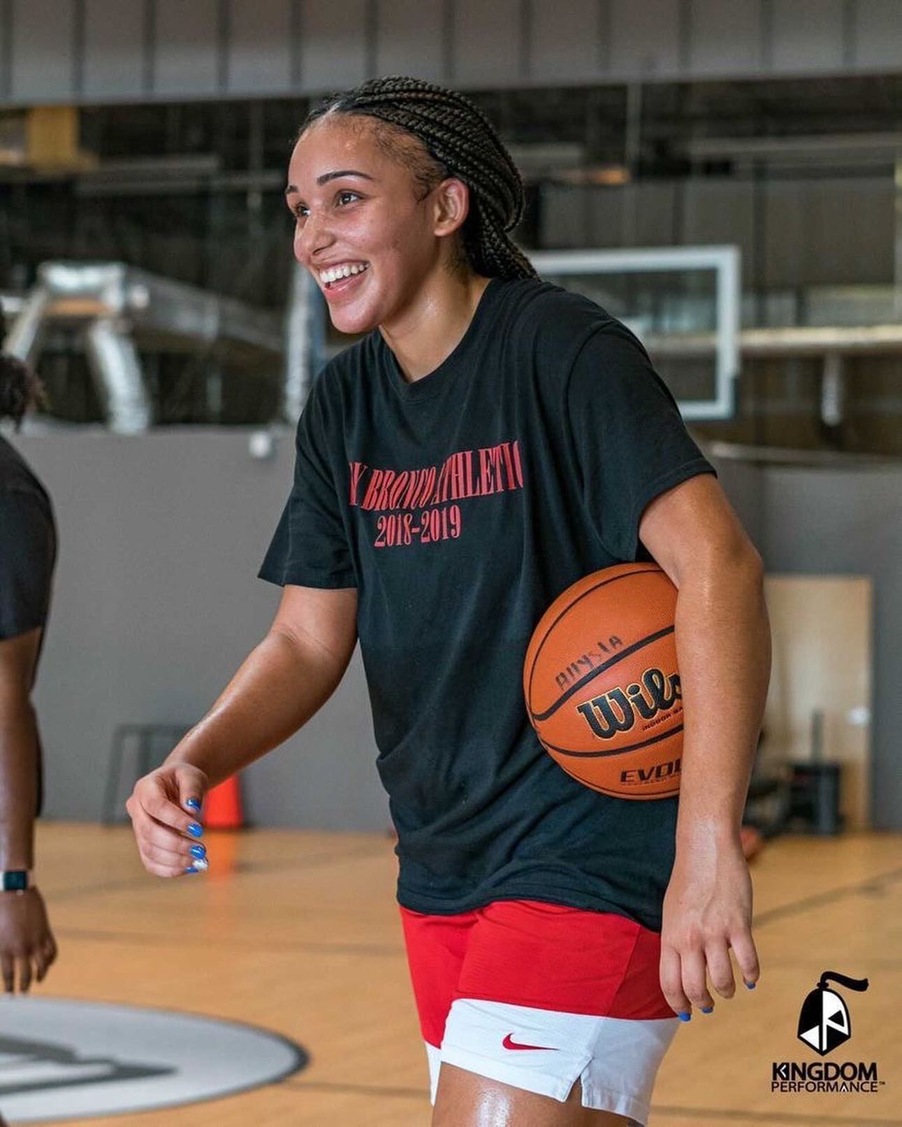 Basketball player in red shorts and black shirt smiles, holding a ball, in a gym.