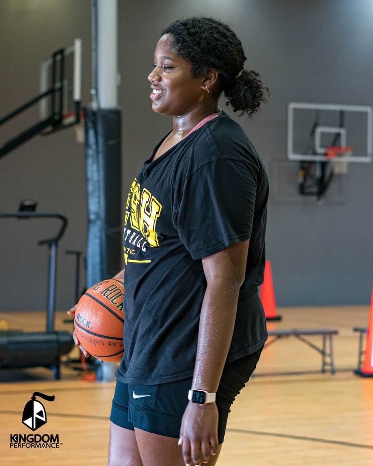 Woman in black shorts and tee holds basketball in a gym.
