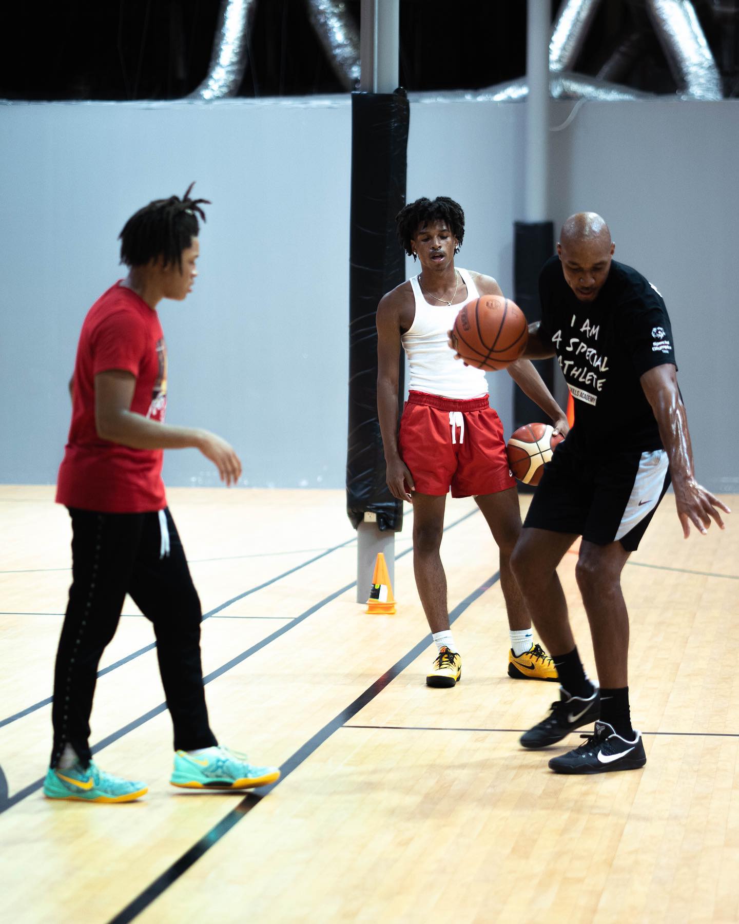 Basketball coach training two teens indoors. Coach in black, teens in red and white, holding a ball.