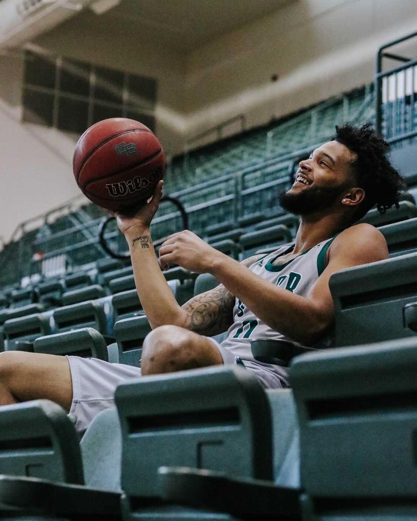 Basketball player smiles holding the ball in the stands; wearing a grey and green jersey.
