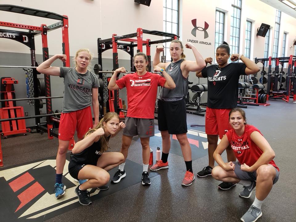 Six women flex their biceps in a gym, wearing athletic gear. Red and black colors dominate the scene.