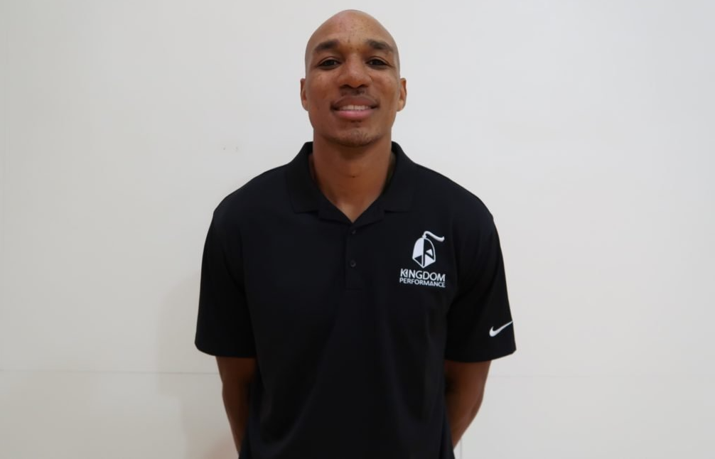 Bald man in black polo shirt with logo, smiling, in front of a plain white wall.