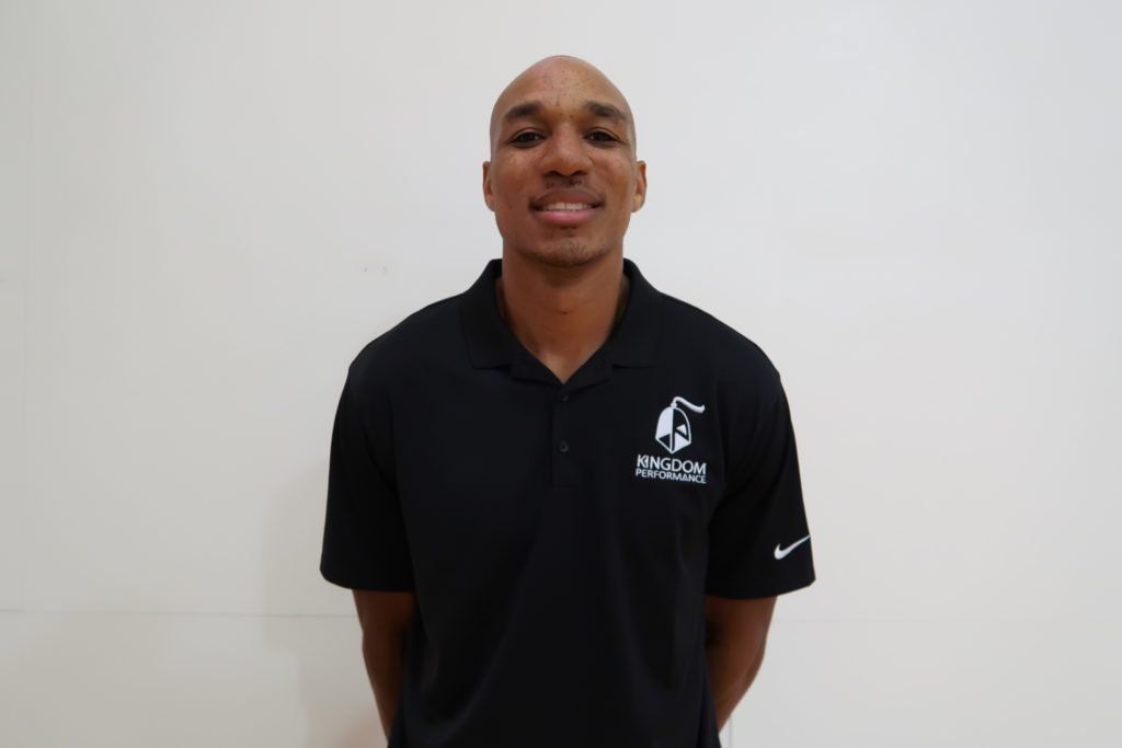 Man in black polo shirt smiles, standing in front of a white wall.
