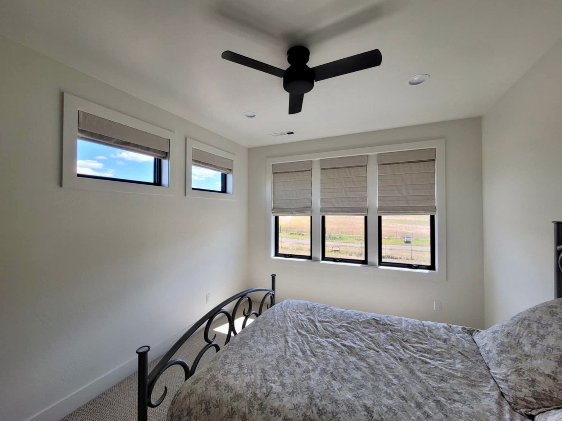 A minimalist bedroom with white walls, a black iron bed frame, a ceiling fan, and windows with neutral Roman shades.