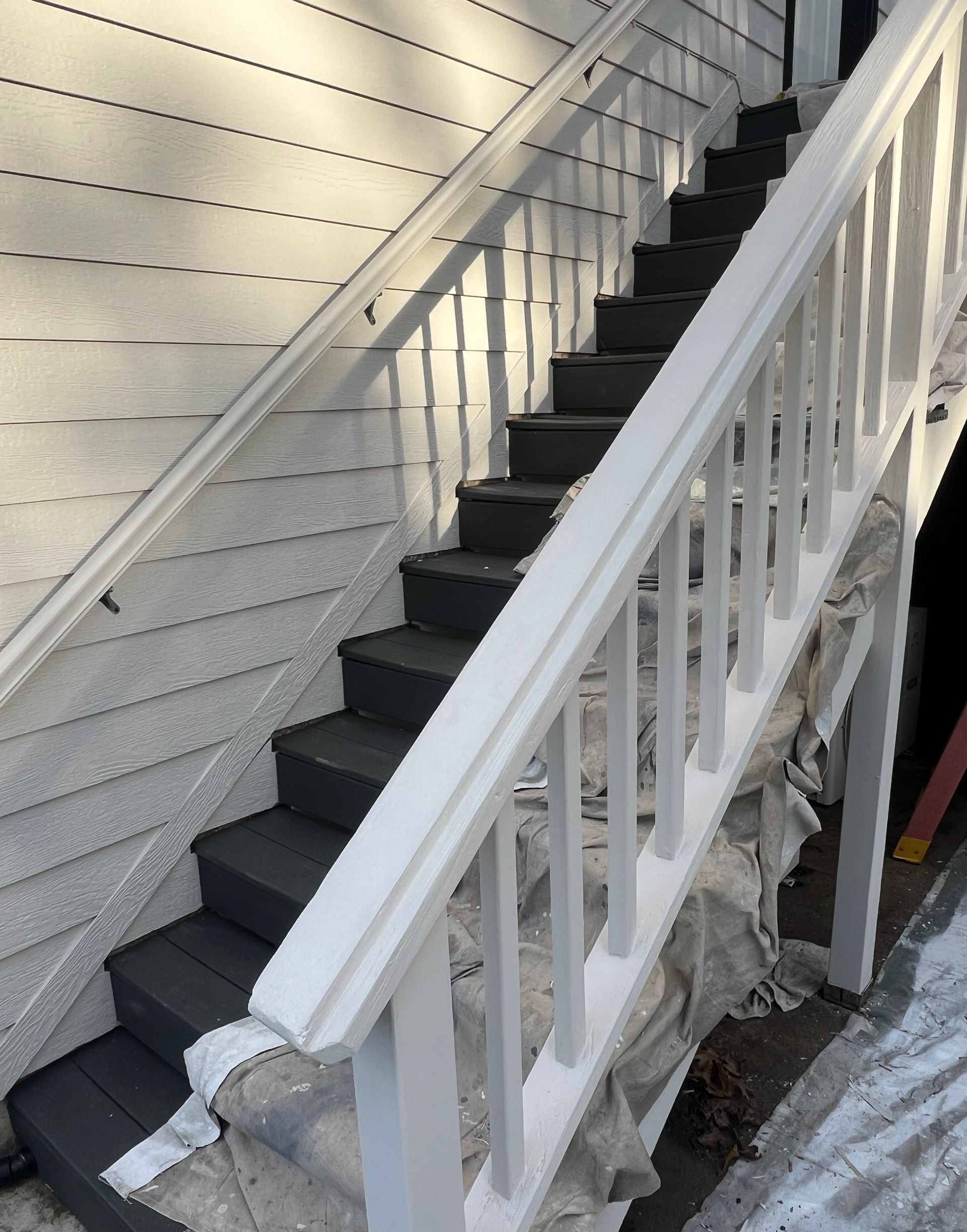 A staircase with dark gray painted treads and white railings against white siding, with a drop cloth underneath.