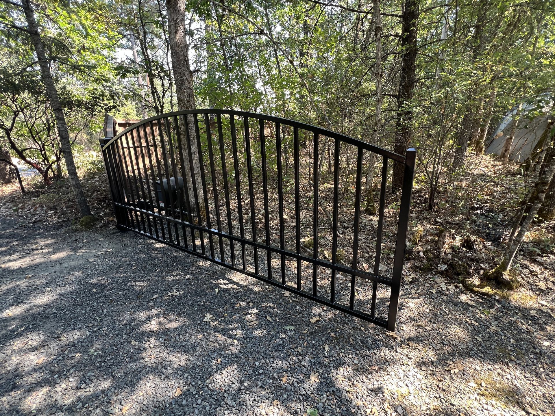 An arched black metal driveway gate stands closed on a gravel path, surrounded by trees and brush.