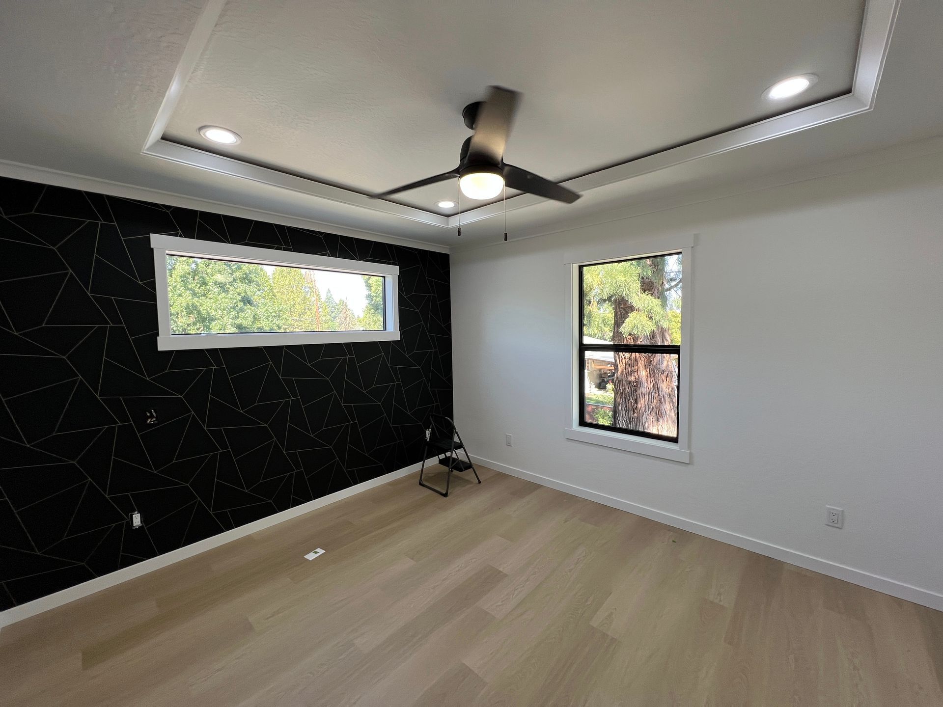 Empty room with light wood floors, a black accent wall with white geometric patterns, a ceiling fan, and two windows.