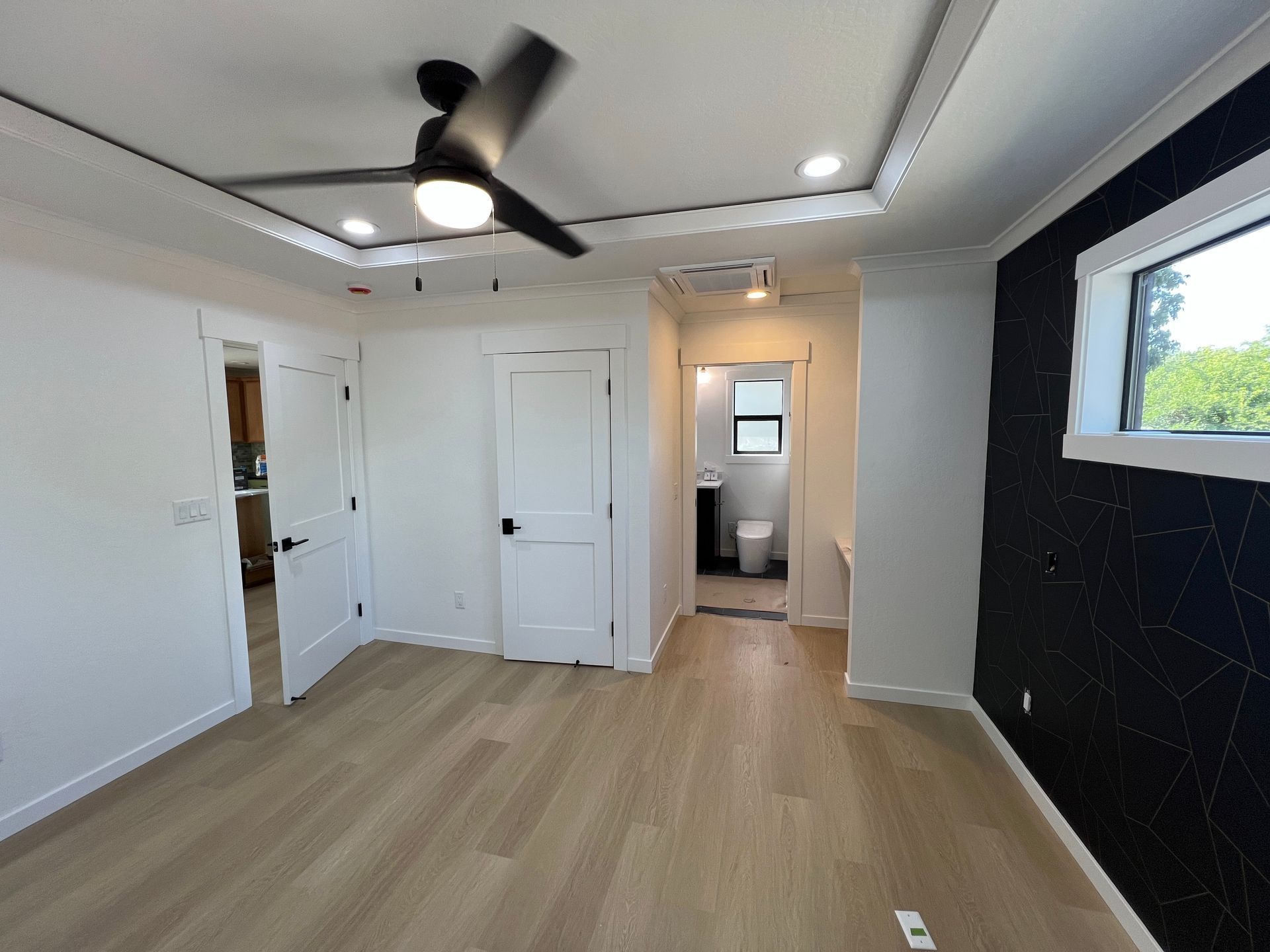 A bright bedroom with wood flooring, white walls, a ceiling fan, and a black accent wall with a window.