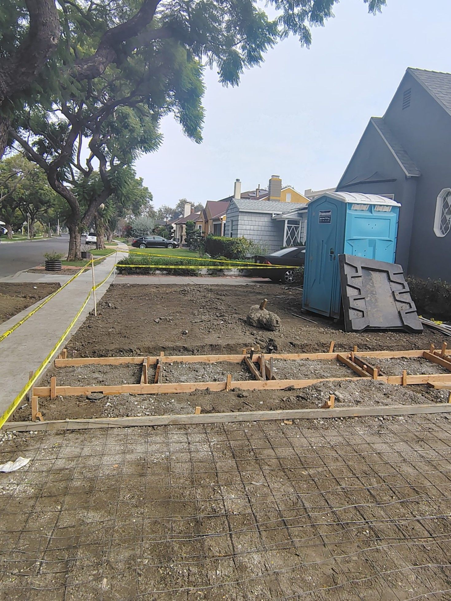 Sidewalk and street under construction beside blue portable toilets, with trees and houses in the background.