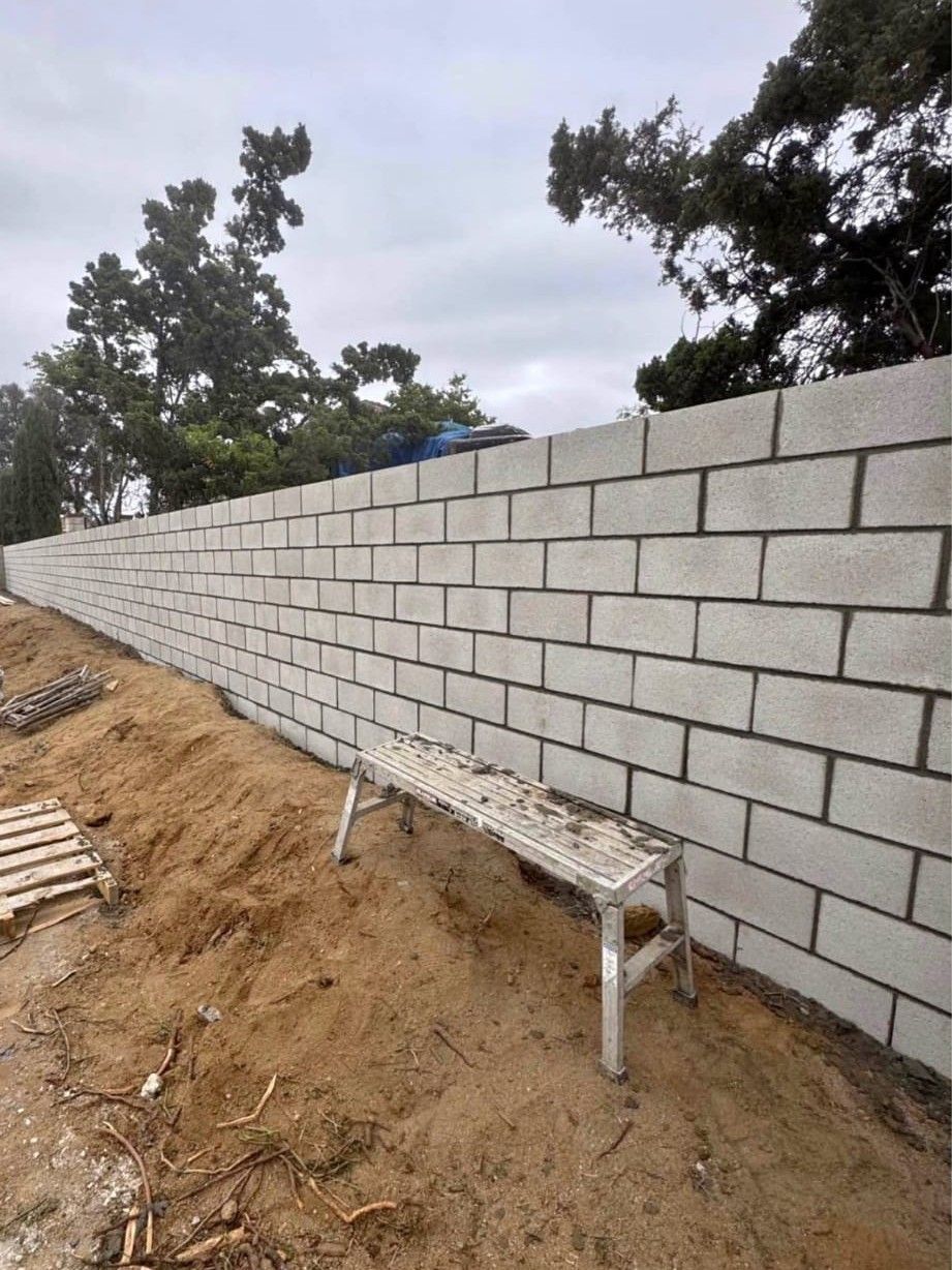 Long concrete block wall under construction along a dirt slope with trees and a bench in front