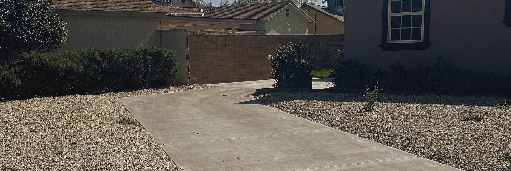 Concrete walkway leading to a house entrance, bordered by gravel landscaping and shrubs in bright sunlight