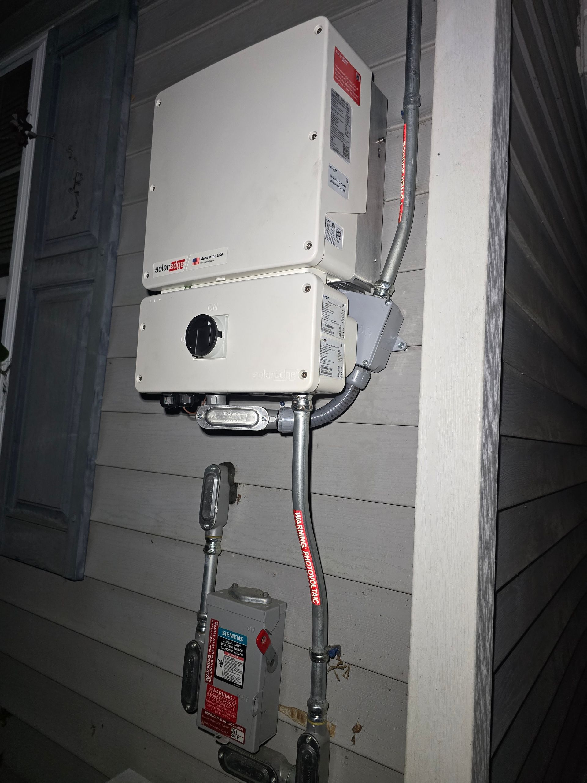 Solar power inverter and electrical components mounted on a house exterior. Gray siding, white boxes, and metal conduit visible.