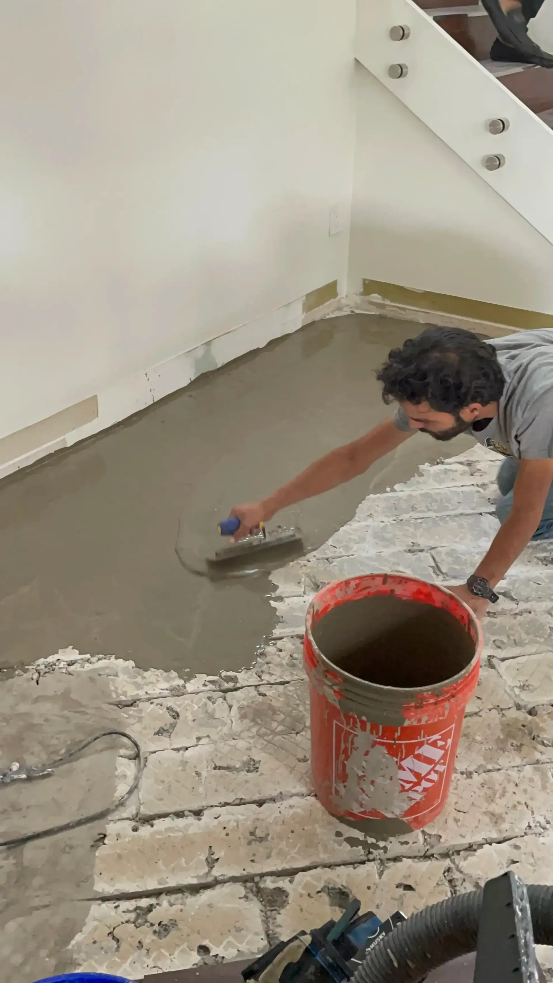 Worker smoothing wet concrete floor with a trowel beside a red bucket in a stairwell.