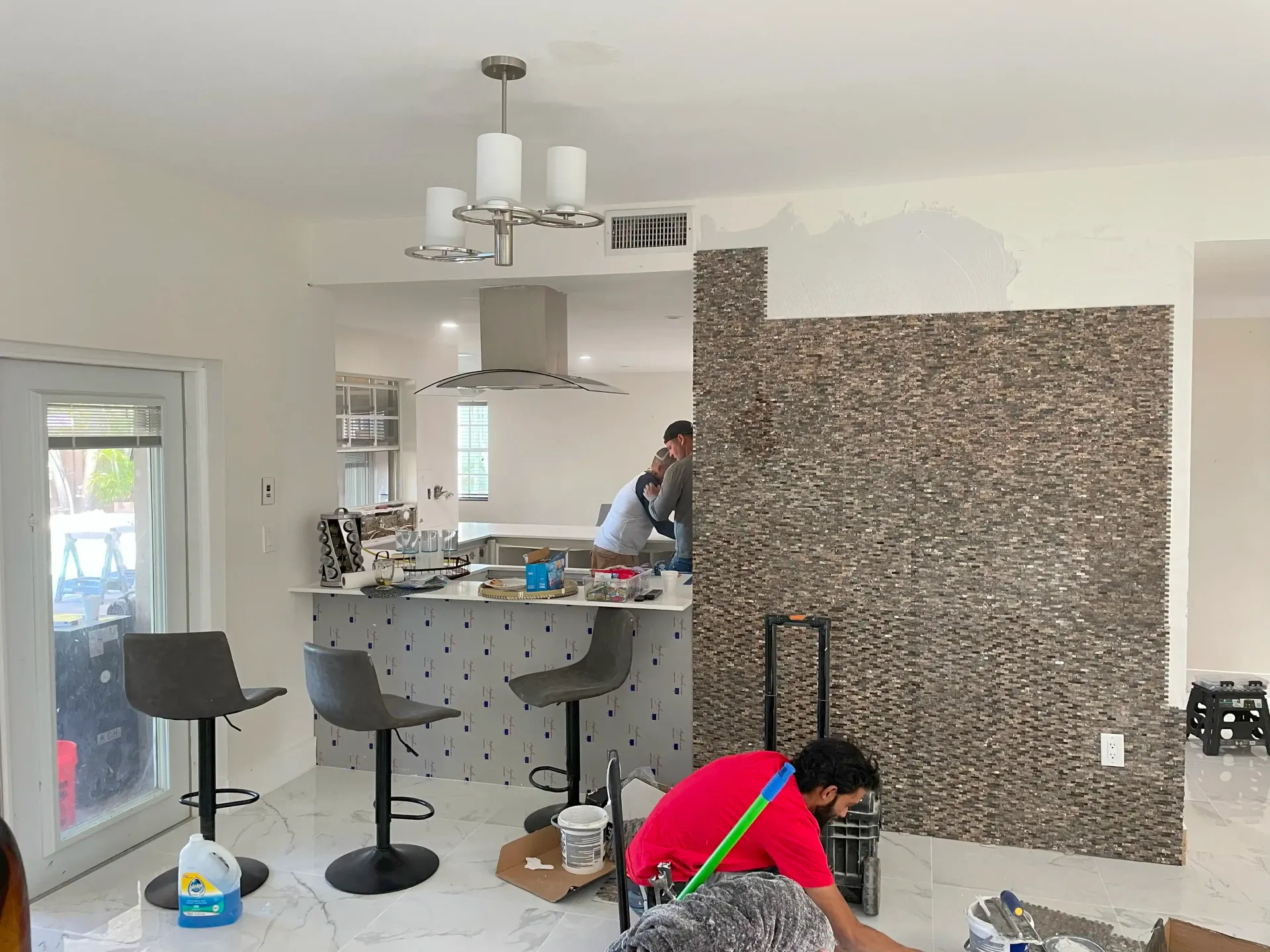 Kitchen with a worker installing a large brown backsplash, with tools and two barstools nearby.