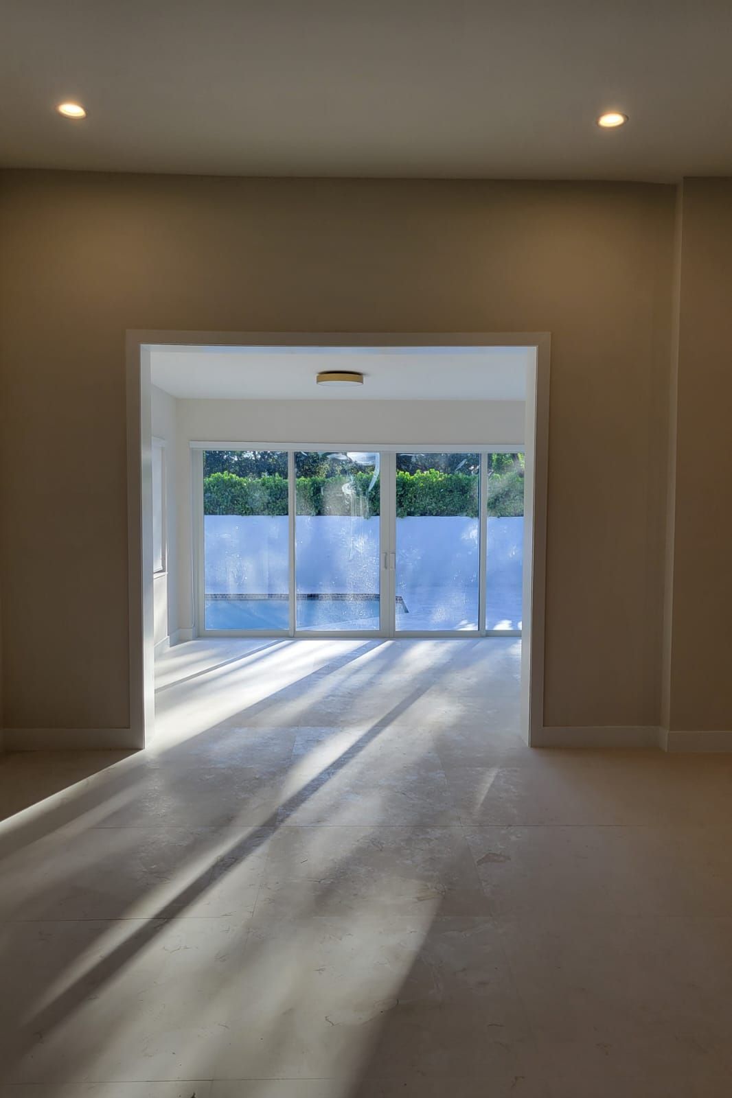 Empty room with beige walls and floor, looking out through open sliding glass doors to a sunlit patio.