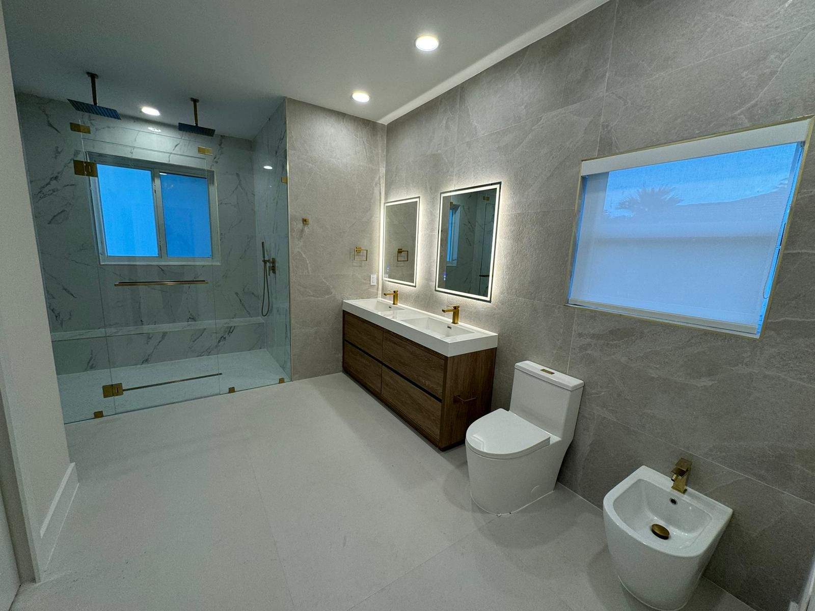 Modern bathroom with glass shower, double vanity, toilet, and bidet in gray tones.