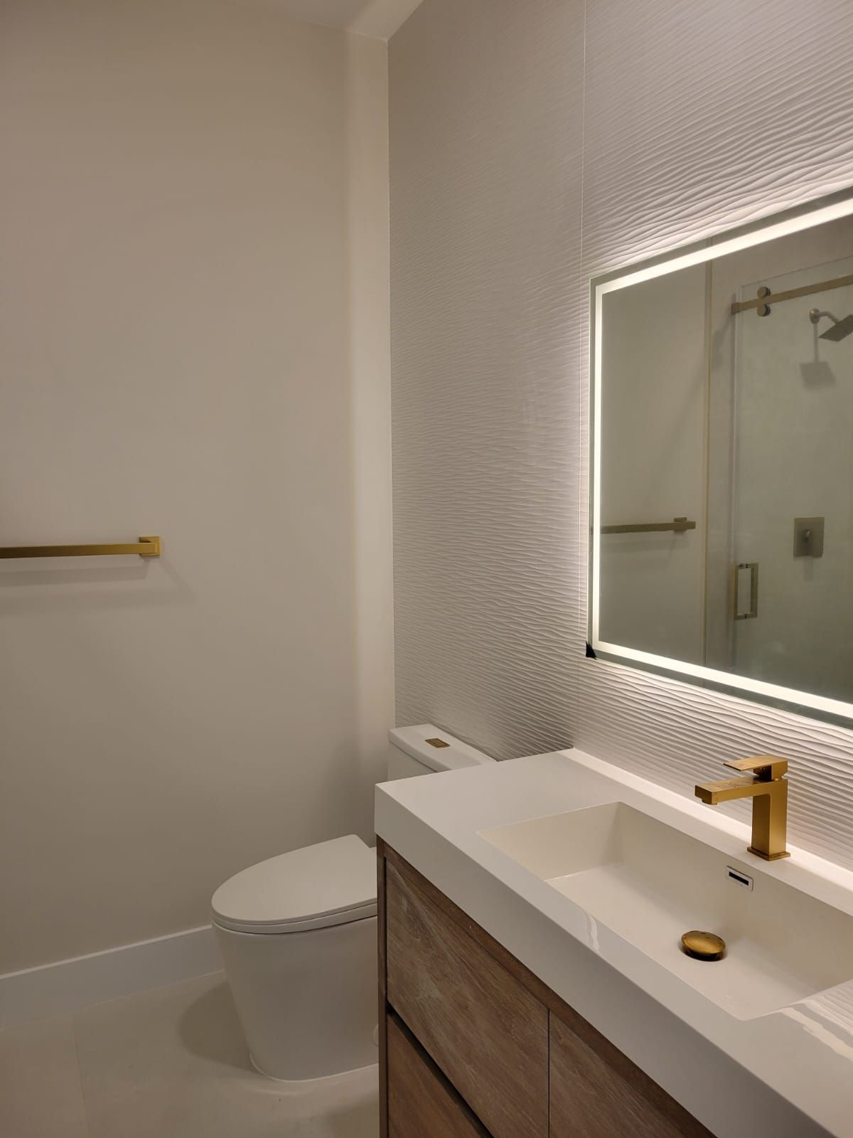 Modern bathroom with toilet, floating wood vanity, gold fixtures, and lit mirror