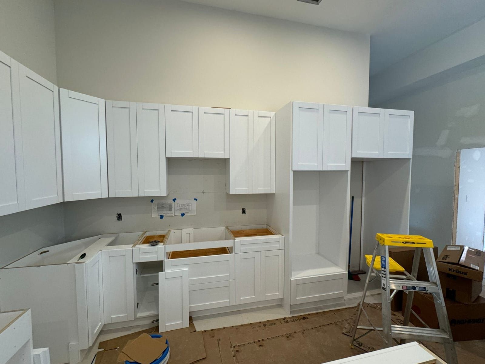 Unfinished white kitchen with cabinets, countertop boxes, and a yellow step ladder on a plastic-covered floor
