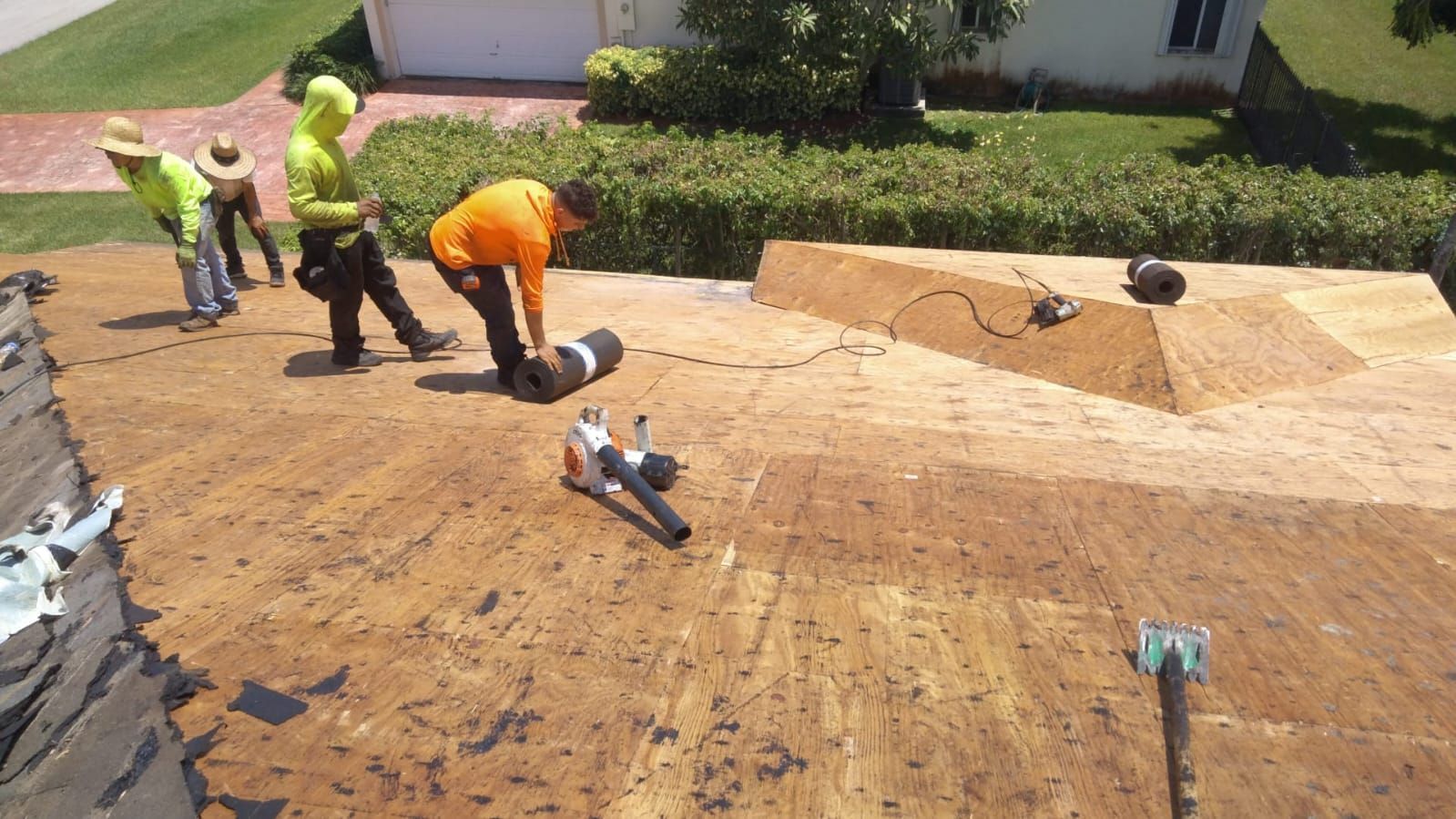 Workers installing roofing shingles on a steep roof, with tools and materials spread across the surface.