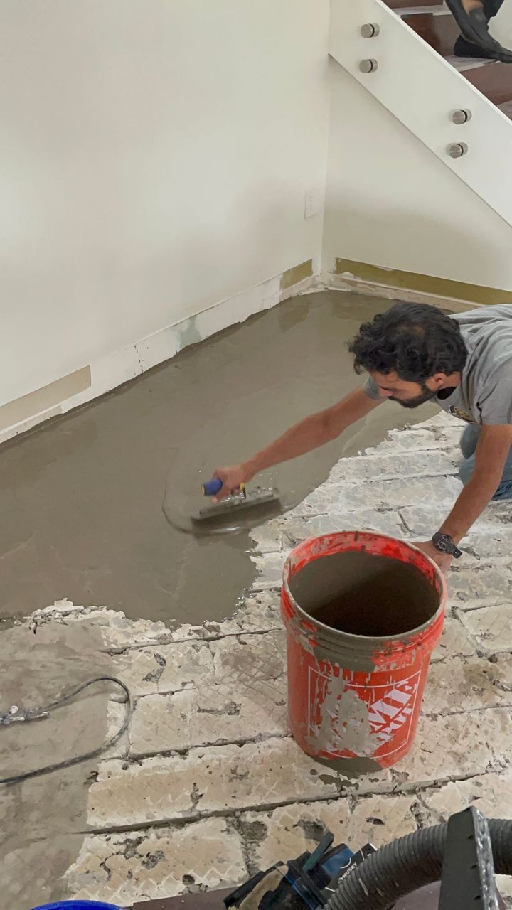 Worker smoothing wet concrete floor with trowel beside an orange bucket in a room under renovation