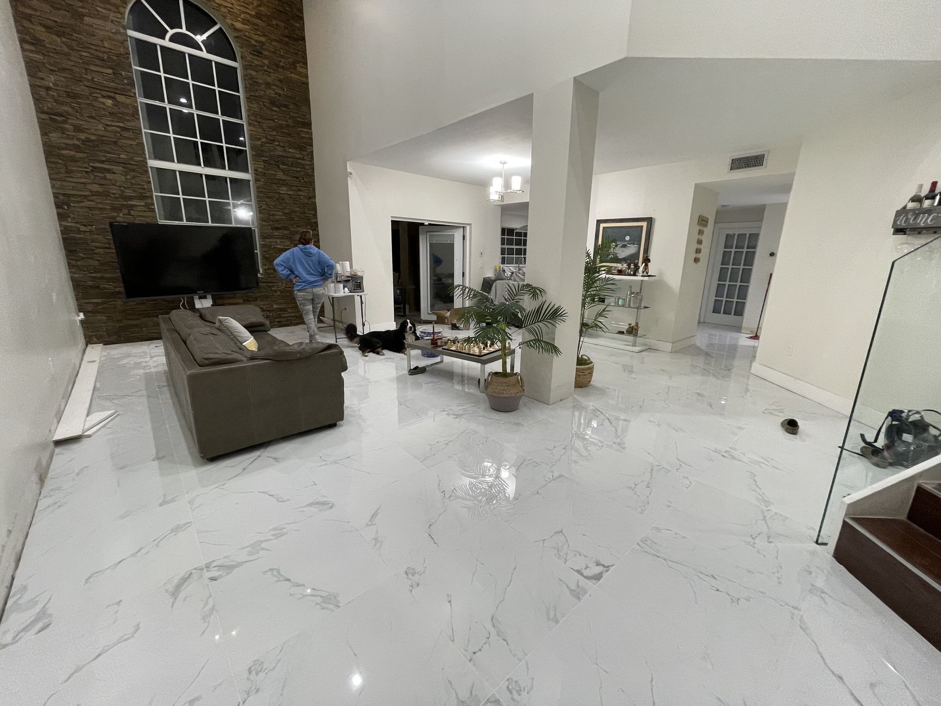 Modern living room with white marble floor, gray sofa, plants, and a person cleaning near the window