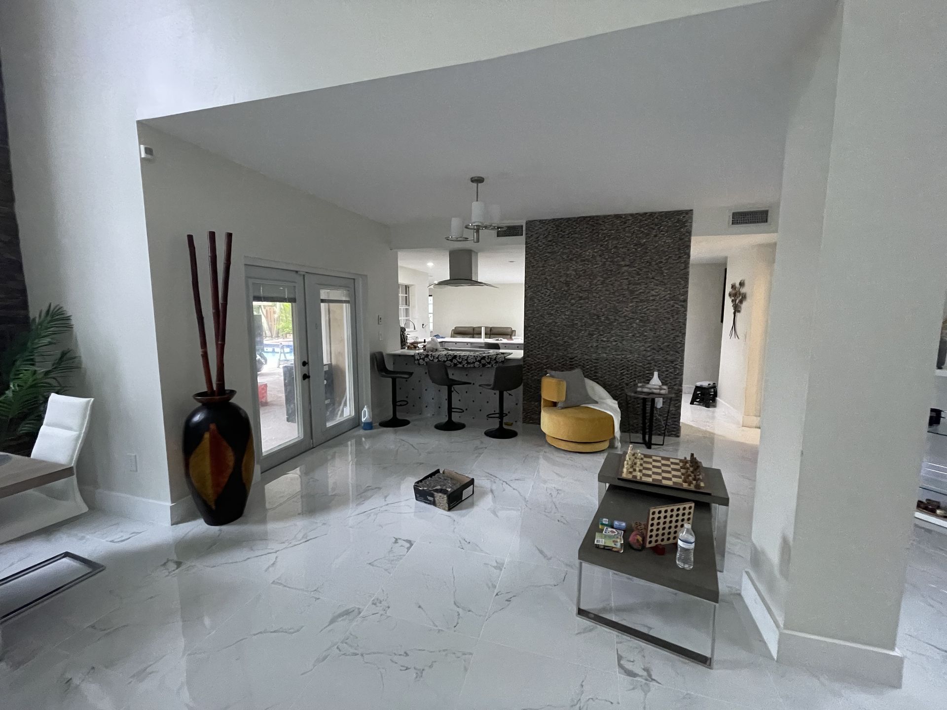 Modern open-plan living room with white tile floors, kitchen island, dark accent wall, and yellow chair.