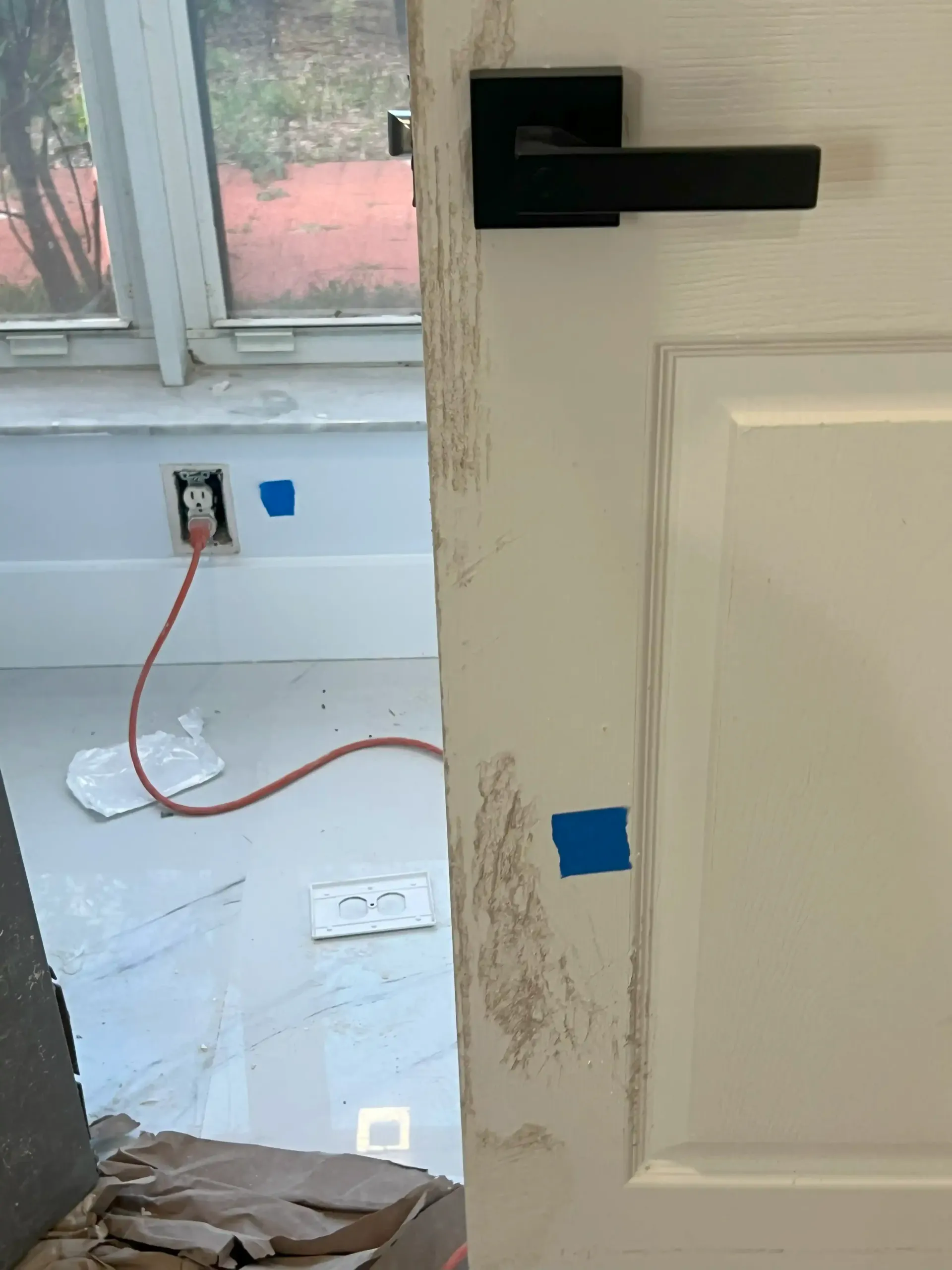 Partially open white door with black handle beside a messy room and window, with blue tape on the wall and outlet