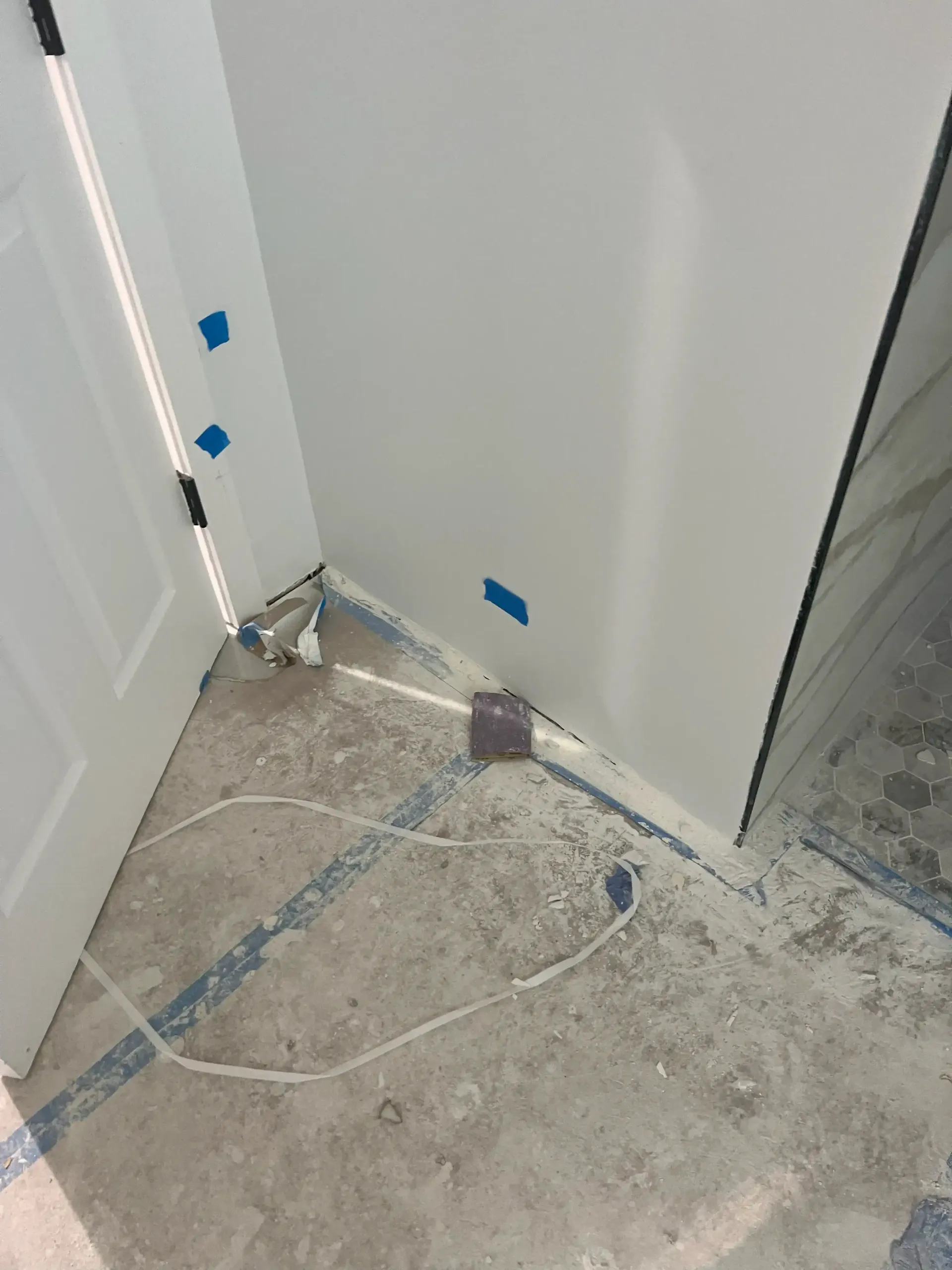 Partially painted corner with blue tape, white wall, and dusty concrete floor near a door and threshold