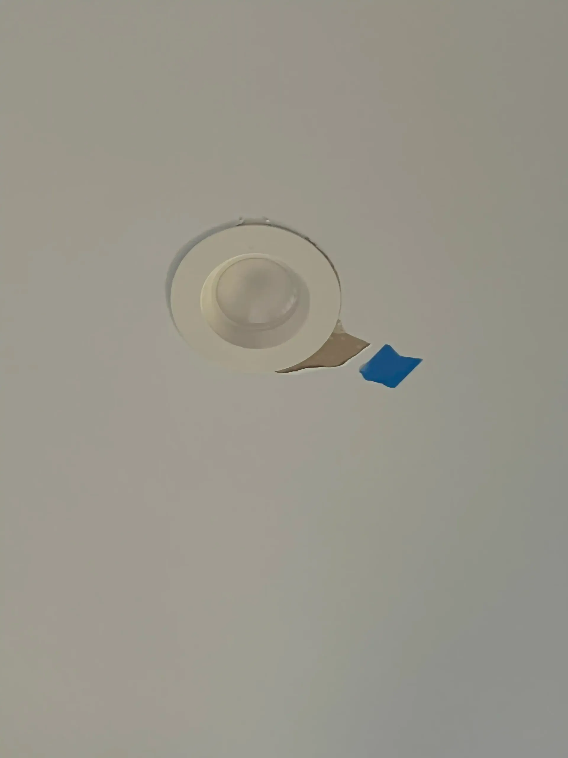 Ceiling smoke detector with a piece of blue tape attached on a plain off-white ceiling