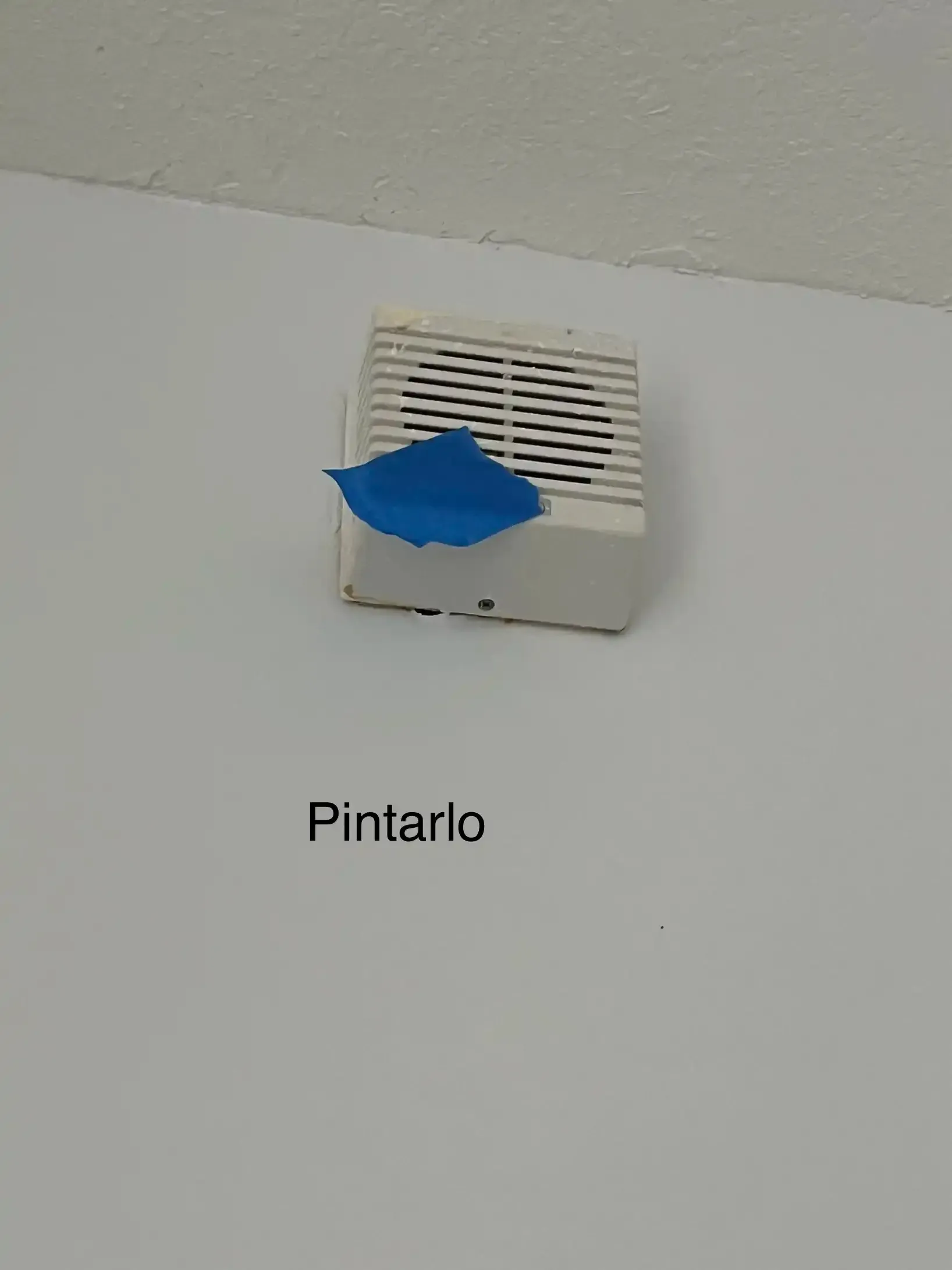 Blue tape covers a wall vent above the word “Pintarlo” on a white wall.