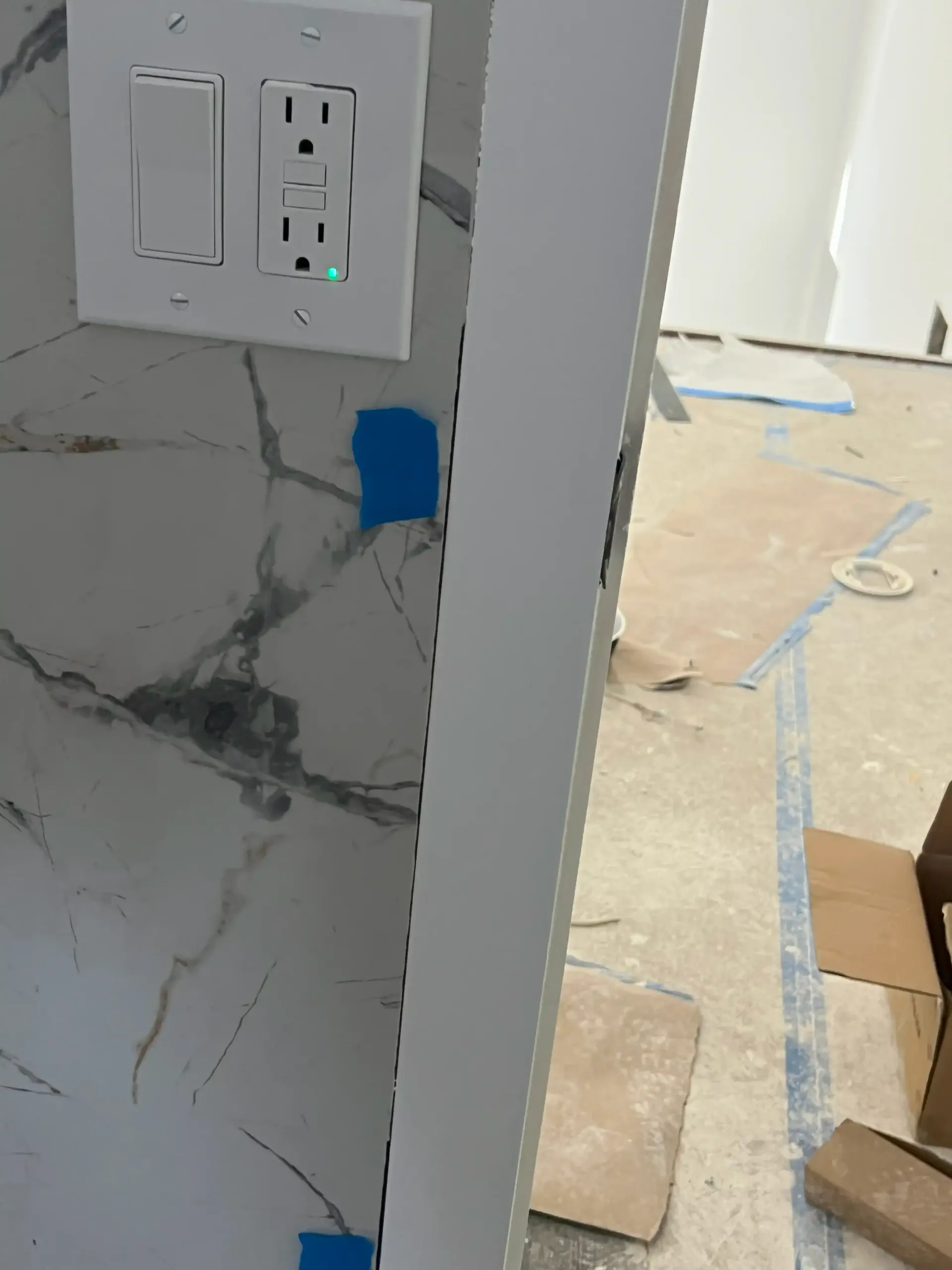 Partially finished kitchen doorway with marble-pattern wall, outlets, blue painter’s tape, and flooring tools below
