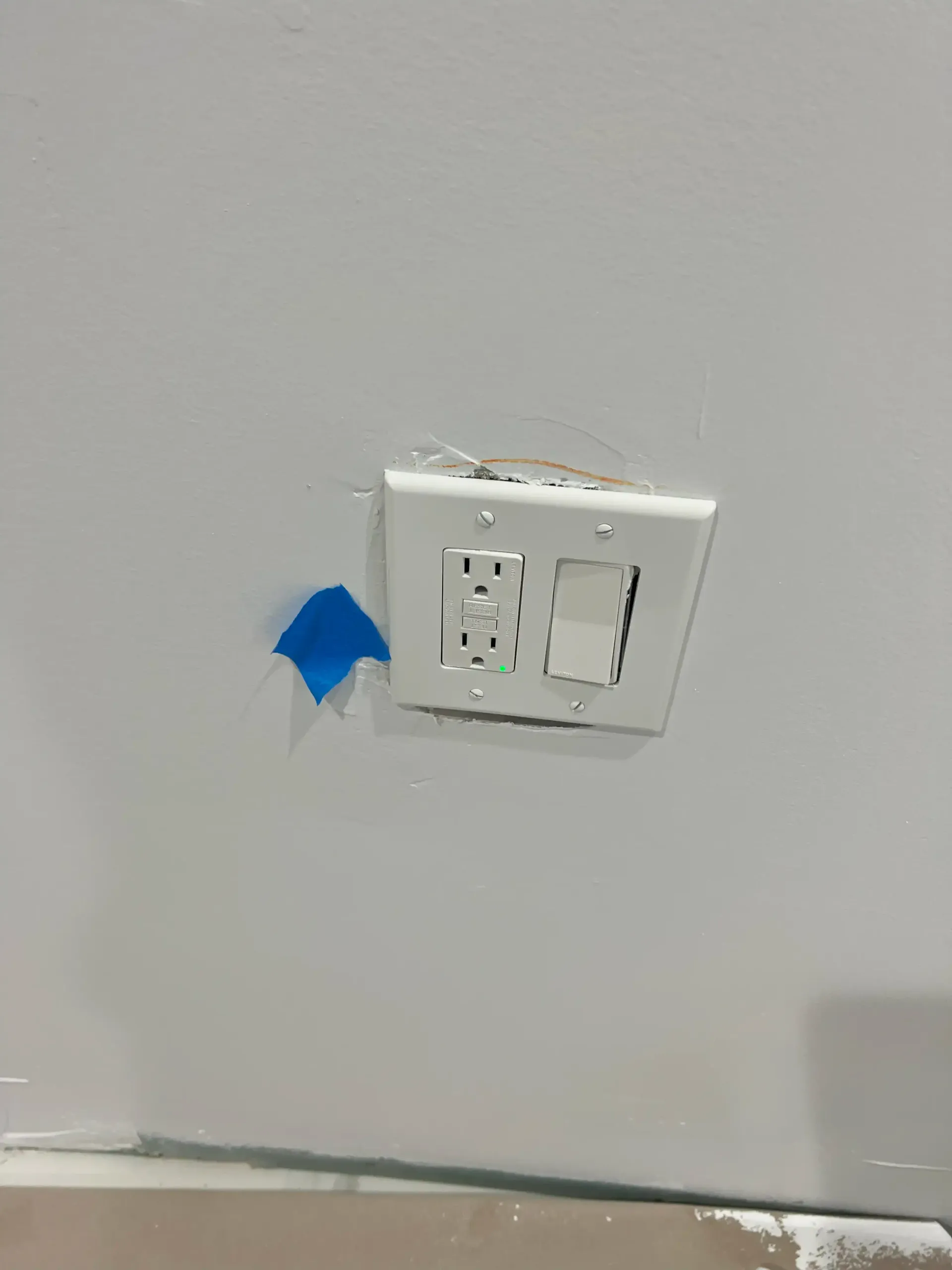 White wall with electrical outlet and light switch plate, marked with blue painter’s tape nearby