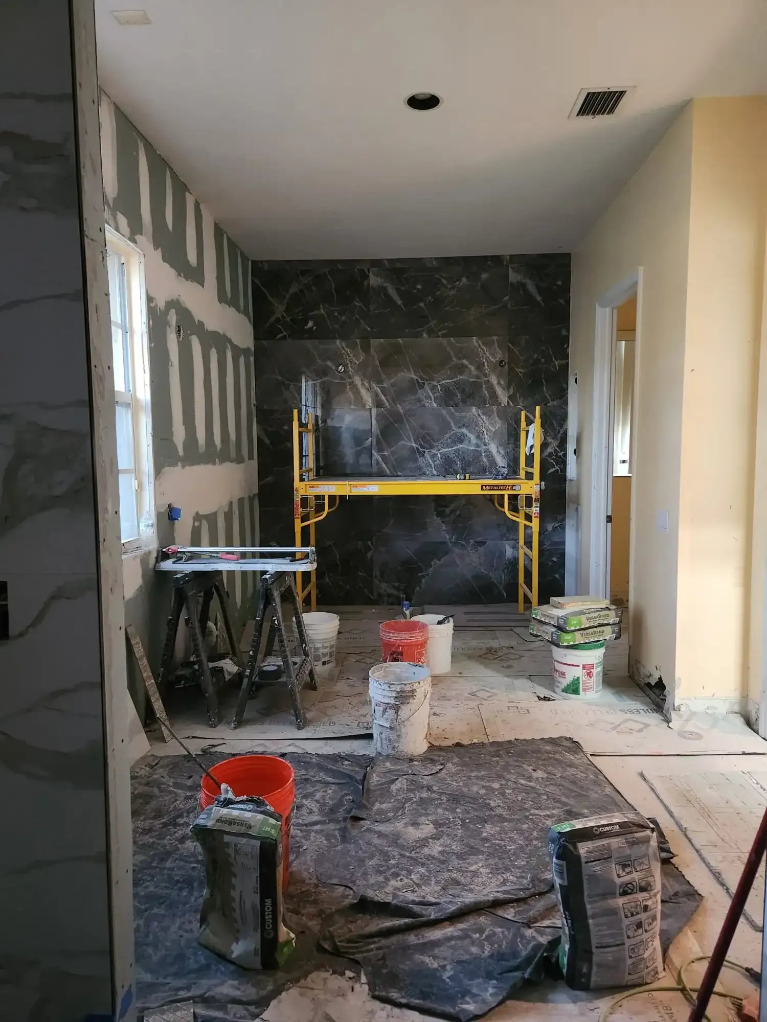Narrow hallway under renovation with drywall, scaffolding, buckets, and a dark tiled accent wall at the end