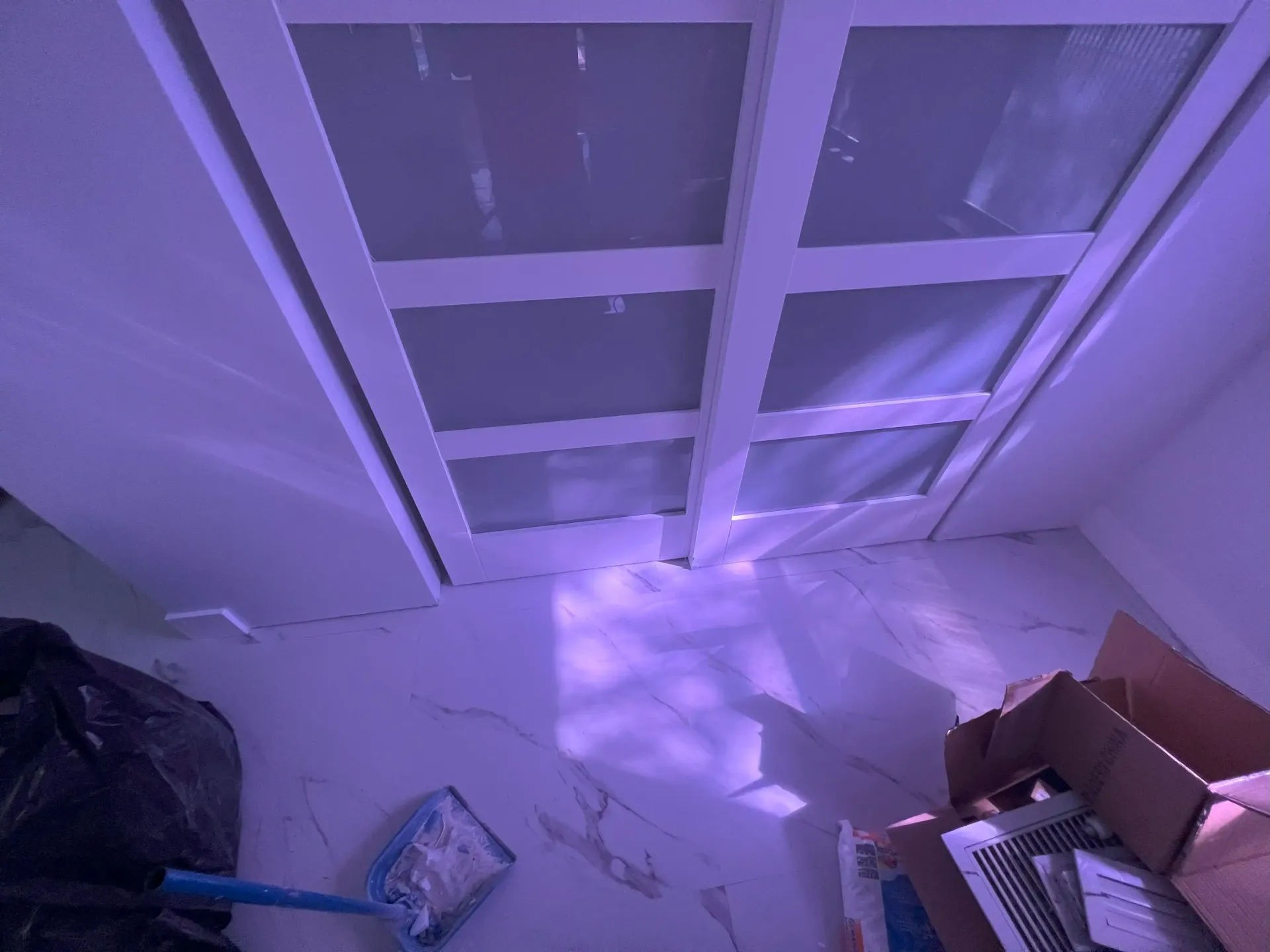Purple-lit closet with open cardboard boxes, a broom, and a black trash bag on the floor