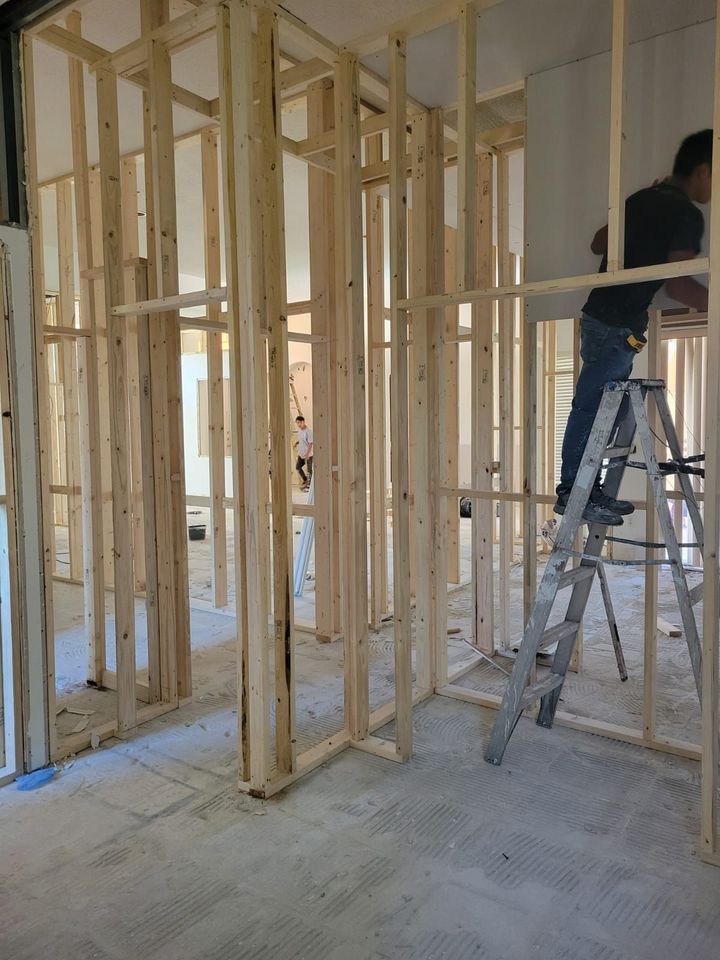 Unfinished room with exposed wooden framing and a worker on a ladder near a partially framed wall