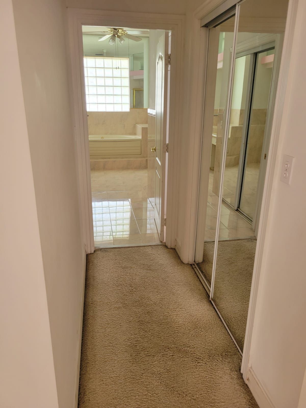 Narrow beige hallway with mirrored closet doors leading to a bright bathroom or bedroom.