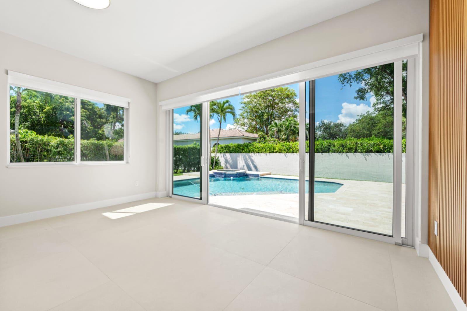 Bright empty room with white tile floors and sliding glass doors opening to a pool and garden.