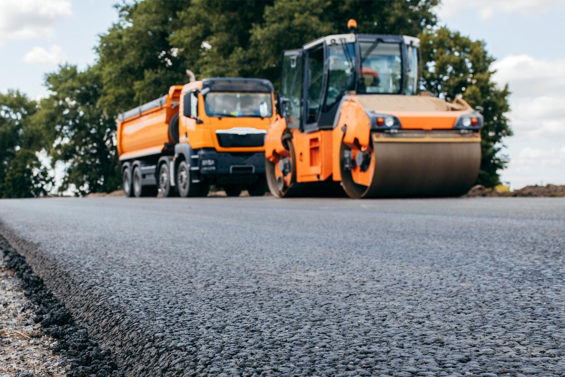 Orange road roller and dump truck on freshly paved road under cloudy sky