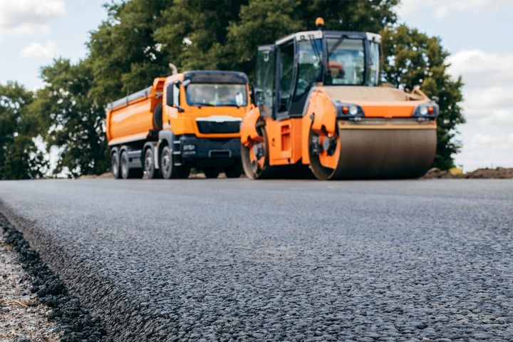 Orange road roller and dump truck on a newly paved road with trees in the background
