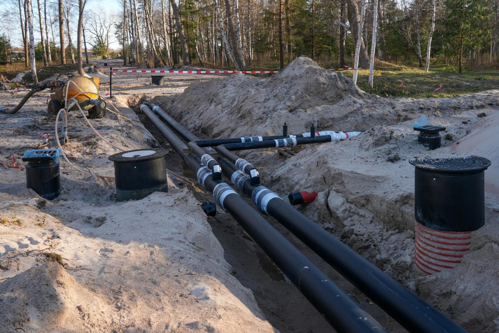 Construction trench with large black pipes and concrete pipes laid in sand at a wooded work site