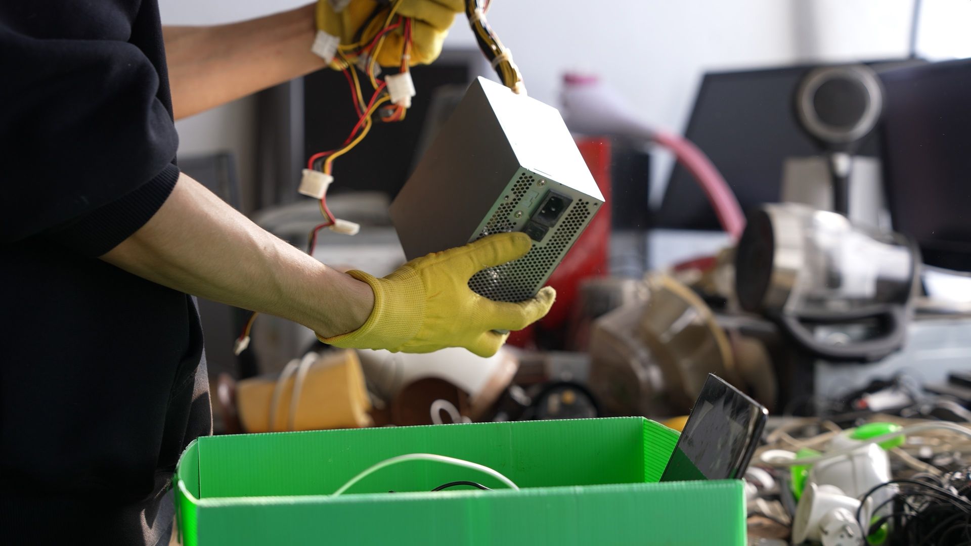Person in yellow gloves placing an electronic device into a green recycling bin at a workbench