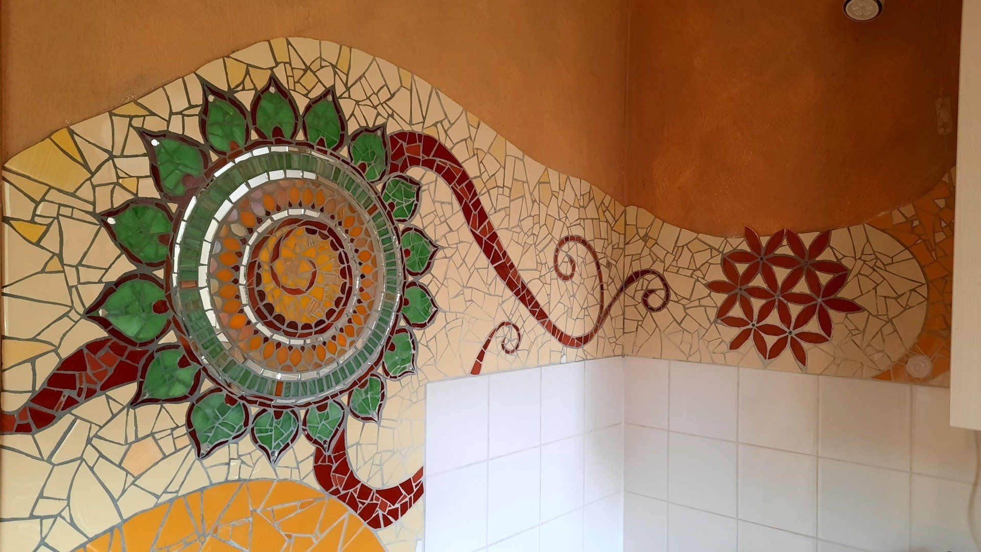 Decoratieve mozaïekmuur met een groene bloem en wervelende rode patronen op een betegelde muur.