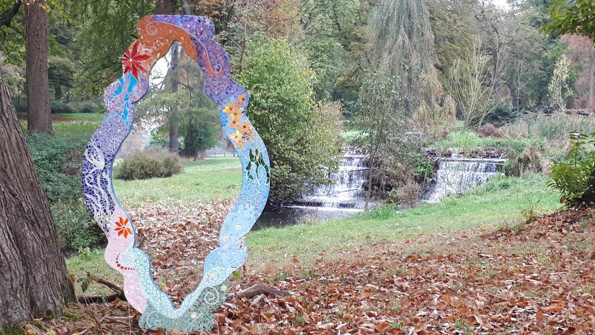Kleurrijke cirkelvormige sculptuur in een park met een waterval en bomen op de achtergrond.