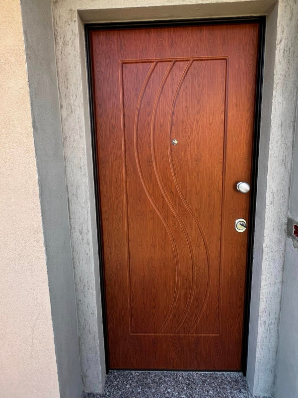 Porta d'ingresso in legno marrone con linee curve decorative, maniglia argentata e spioncino.