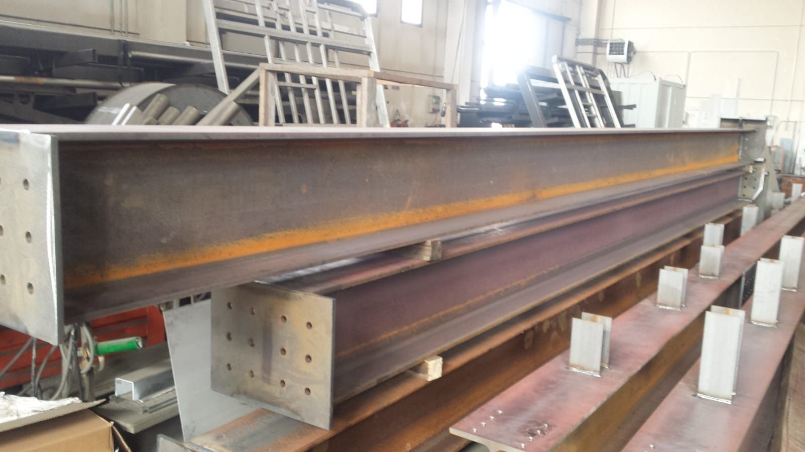 Grandi travi a I in acciaio con piastre terminali imbullonate, appoggiate su supporti in un'officina di fabbricazione.