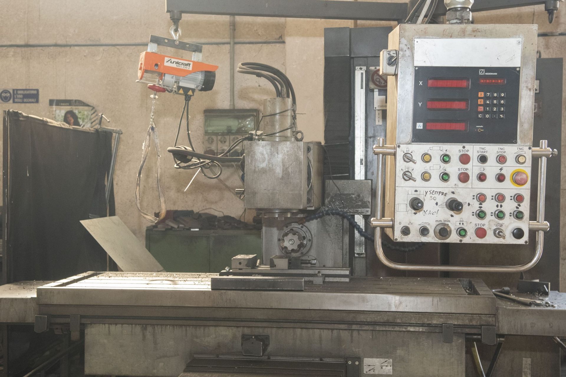 Una grande fresatrice industriale con pannello di controllo, display digitale e paranco sospeso sopra l'area di lavoro.